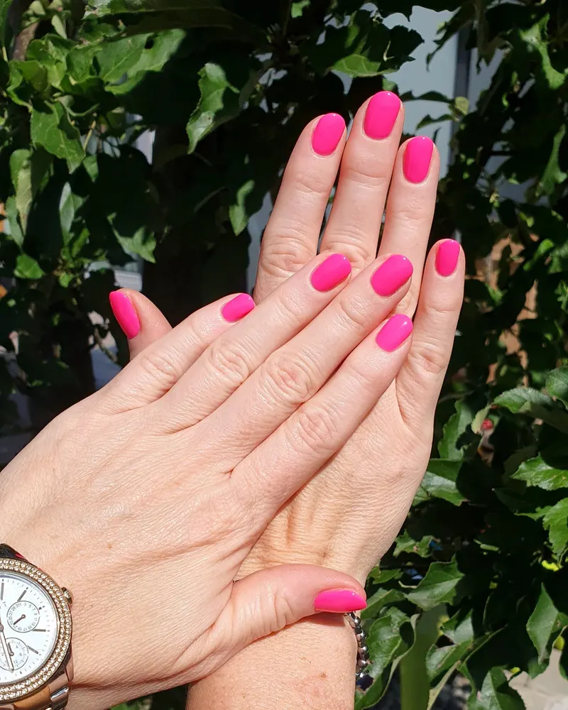 Kräftig pinke Nägel, kurz und rund, vor grünem Laub