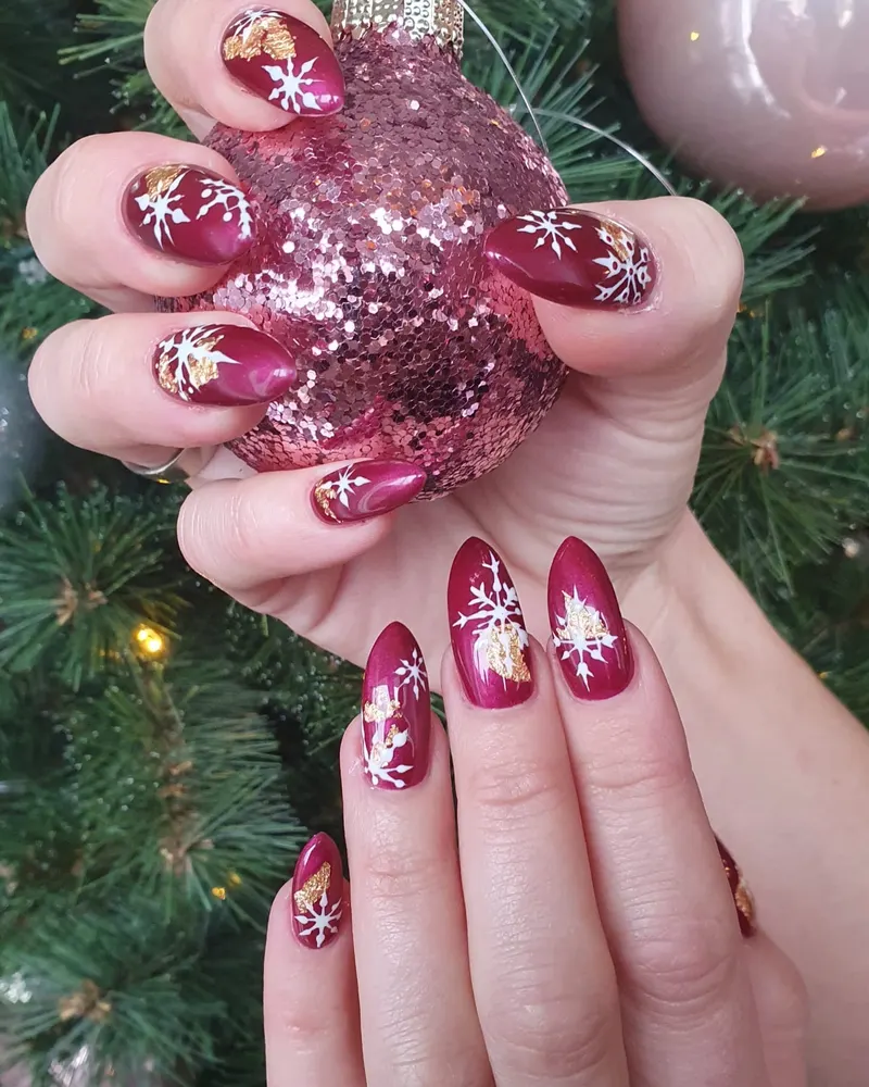 Bordeauxrote Mandelnägel mit weißen Schneeflocken neben einer glitzernden Weihnachtskugel