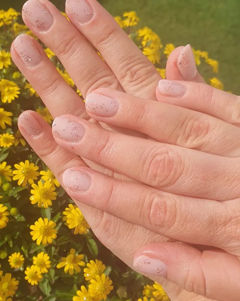 Nude-Nägel mit feinen Glitzerpunkten vor gelben Blüten